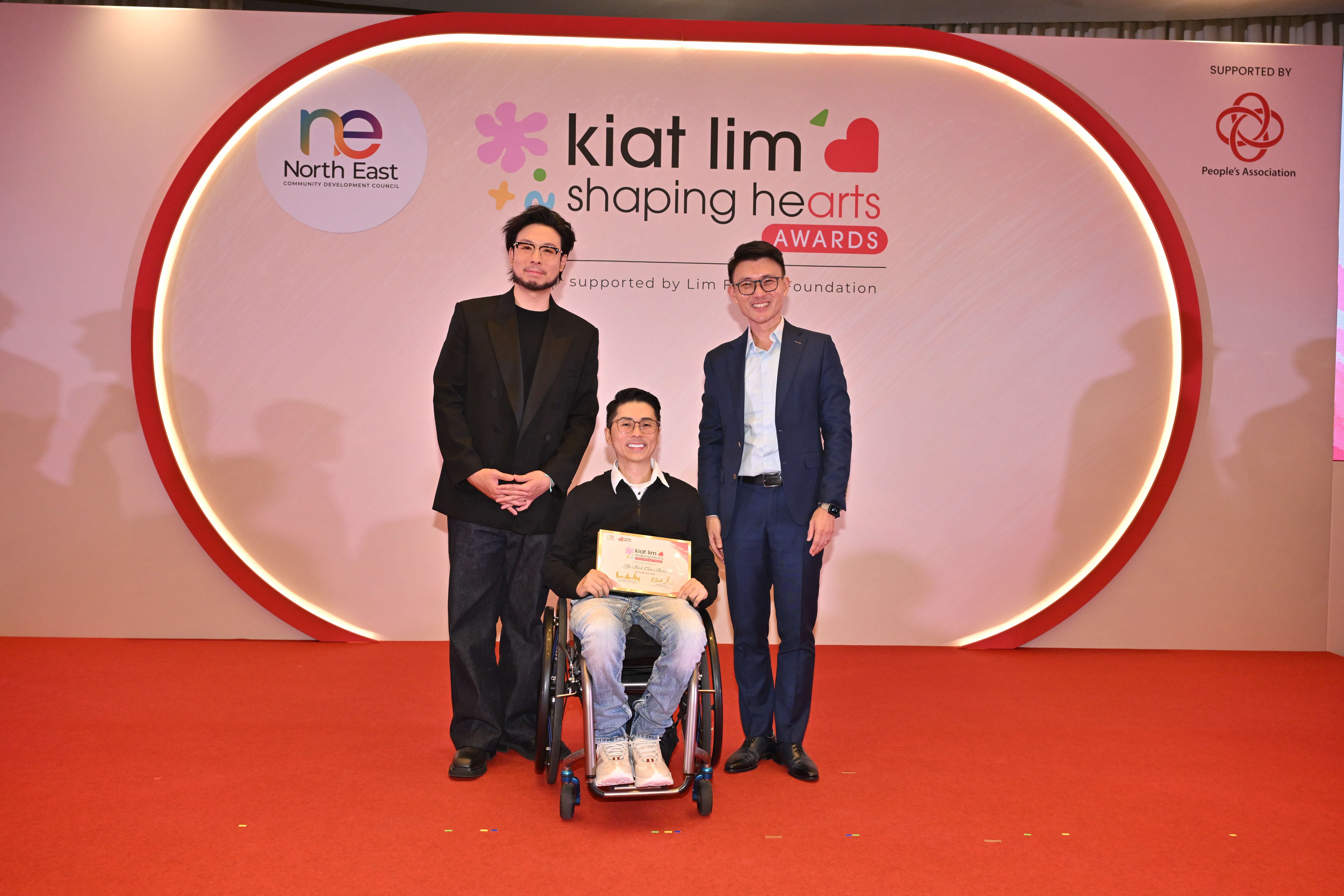 Award recipient standing on stage with the presenter, holding their certificate and posing for a group photo during the award ceremony, with a backdrop reading ‘Kiat Lim-Shaping Hearts Awards'.